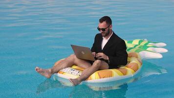 Businessman in a suit sitting on a pineapple-shaped inflatable, working on a laptop in a pool. Concept remote work, summer. video