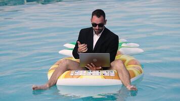 Businessman in a suit sitting on a pineapple-shaped inflatable, working on a laptop in a pool. video