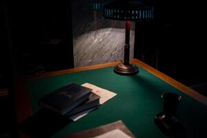 A Desk With a Green Felt Surface Illuminated by a Vintage Lamp Showcases Books and Papers in a Dimly Lit Room During the Night photo