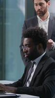Caucasian leader boss come to African American employee multicultural diversity businessmen men males coworkers colleagues working computer tablet discussing business project teamwork guys talk office video