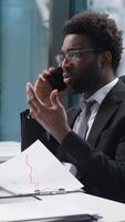 Confused African American man employee talking mobile phone businessman business cellphone looking computer monitor pc worker manager male call problem trouble puzzled guy consulting client in office video
