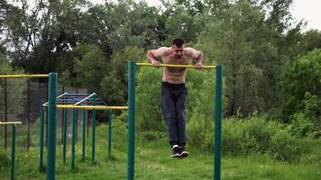 Strong and muscular man doig exercises on horizontal bar at nature. Hardy shirtless sportsman working out at sports ground. Young athletic guy exercising outdoor. Concept of healthy lifestyle video