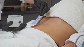 cosmetologist uses spatula or applies cream to patient's stomach in preparation for epilation procedure. patient, young woman, is lying on couch. video