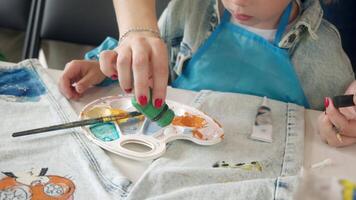 A young mother is engaged in creative work with her child, carefully decorating denim clothes together using bright watercolor paints. video