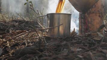 Pouring hot drink into metal mug at forest campsite, Steaming beverage flows into tin cup on forest floor near rustic fire setup during outdoor survival video
