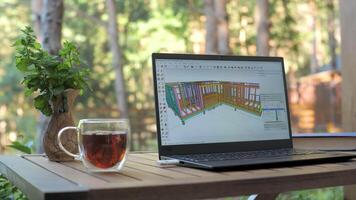 Architectural framing design on laptop in natural environment, Detailed wooden frame model displayed on screen in forest setting with tea and potted herb video