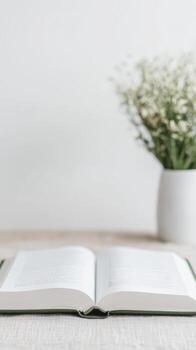 An open book rests on a light wooden surface, beside a vase of delicate white flowers, embodying a serene and minimalist scene of quiet study and peaceful contemplation. photo
