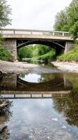 Scenic concrete bridge arches gracefully over a crystal-clear river, reflecting lush green trees and serene skies, a peaceful natural landscape. photo