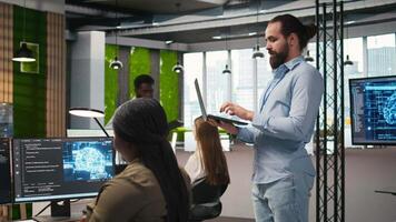 Jolly programmer coding AI algorithms on laptop to debug software issues and improving system stability. Smiling employee in tech startup office configuring settings using notebook, camera A video