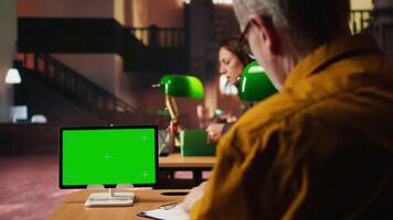 Elderly adult in a public library studying for thesis next to copy space display, preparing paperwork under a green lamp at desk. Old man surrounded by textbooks and study materials. Camera A. video