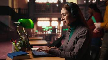 Mature student listening to school webinar on laptop at a library, takes notes and does research during online lecture. Studying remotely with an expert to obtain her diploma. Camera A. video