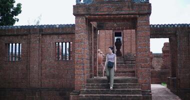 Handheld rear view, Relaxed young woman in a striped shirt and green pants walks around, admiring old sculptures and ancient brick structures, Enjoy holiday travel. video