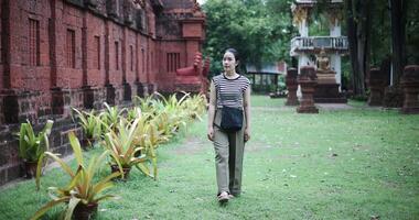 Handheld shot, Relaxed young woman wearing a striped shirt and green pants walks around, admiring the natural scenery and ancient brick structures, Enjoy holiday travel. video