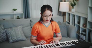 Handheld high angle view shot, Young Asian woman wearing glasses and an orange t-shirt is playing the keyboard with a smile in a beautifully decorated living room. video