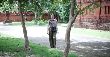 Handheld shot, Relaxed young woman wearing a striped shirt and green pants walking around, admiring the natural scenery and ancient brick structures, Enjoy holiday travel. video
