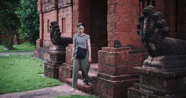 Handheld shot, Relaxed young woman in a striped shirt and green pants walks around, admiring old sculptures and ancient brick structures, Enjoy holiday travel. video