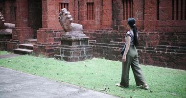Rear view shot, Relaxed young woman in a striped shirt and green pants walks around, admiring old sculptures and ancient brick structures, Enjoy holiday travel. video