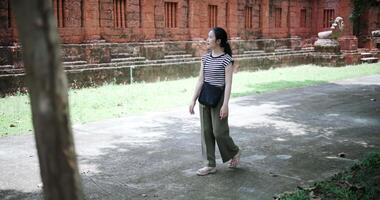 Relaxed Asian woman wearing a striped shirt and green pants walking around, admiring the natural scenery and ancient brick structures video