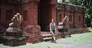 Relaxed young woman in a striped shirt and green pants walks around, admiring old sculptures and ancient brick structures, Enjoy holiday travel. video