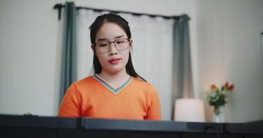 Handheld low angle view shot, Young Asian woman wearing glasses and an orange t-shirt is playing the keyboard with a smile in a beautifully decorated living room. video