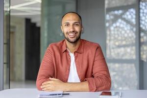 A smiling man sits at a table, appearing confident and relaxed. The setting suggests a modern office or workspace. photo