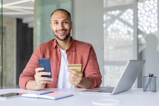 A smiling man using a credit card and smartphone, with a laptop and notebook on the table. photo