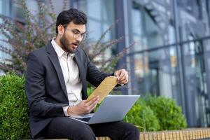 An astonished businessman sits outside with a laptop, examining an envelope with a look of surprise, possibly receiving important documents. photo
