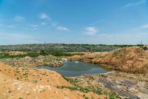 A wasteland of garbage rises in the background with a small pond and patches of greenery under a bright sky photo