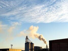 Smoke rises from a factory against a clear sky, highlighting the industrial landscape during the evening hours photo