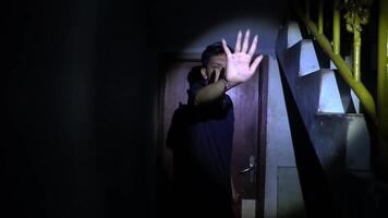 A young man stands in a dimly lit hallway, illuminated by a flashlight. He appears anxious, looking towards a staircase. The atmosphere is tense and suspenseful, suggesting a horror or thriller theme. video