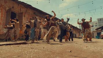 A dynamic dance group performing in an urban setting with rustic buildings and string lights. The dancers wear diverse outfits, showcasing energy and movement in a vibrant atmosphere. video