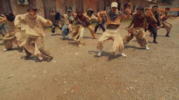 A dynamic group of dancers performing a synchronized routine outdoors, showcasing energetic movements and diverse styles. The setting features rustic buildings and a gravel surface video