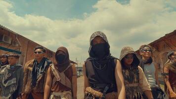 A group of eight individuals dressed in rugged, post-apocalyptic attire, standing confidently in a deserted urban setting. They wear masks and sunglasses, showcasing a mix of styles and accessories video