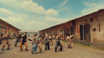 A dynamic group of dancers performing a synchronized routine outdoors, showcasing energy and movement. The setting features rustic buildings and a cloudy sky, enhancing the vibrant atmosphere. video