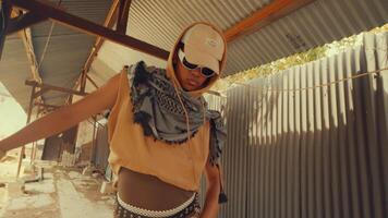 A stylish individual wearing sunglasses and a cap, dressed in a unique outfit with a scarf, stands in an industrial setting with corrugated metal walls and exposed beams. video