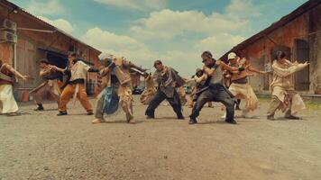 A group of dancers performing a choreographed routine outdoors, set against a backdrop of rustic buildings and a blue sky. The dancers wear a mix of casual and stylized clothing video