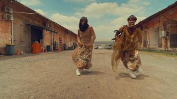 två dansare utför i en rustik utomhus- miljö, bär lösa, mönstrad byxor och blast. de bakgrund funktioner gammal byggnader och en molnig himmel, skapande en vibrerande atmosfär. video