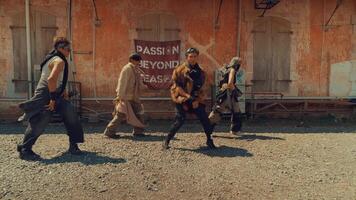 A group of four dancers performing in an outdoor setting with a rustic backdrop. They wear eclectic outfits, showcasing a mix of styles and accessories. The atmosphere is energetic and dynamic video