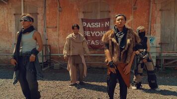 A group of four dancers performing a choreographed routine outdoors, set against a rustic backdrop with an old building and a banner. The dancers wear stylish, urban outfits and display dynamic video