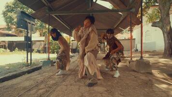 Three dancers performing a synchronized jump in a rustic outdoor setting. The scene captures dynamic movement and energy, with a backdrop of trees and an old building. video