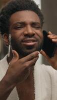 African American man looking at mirror reflection bathroom guy talking mobile phone touching face care shaving beard smiling bearded male call conversation smartphone communication in hotel bath home video