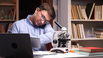 närbild porträtt forskare är arbetssätt sent i laboratorium, använder sig av läsplatta och talande på de telefon, med en mikroskop och böcker på de skrivbord och en bokhylla i de bakgrund video