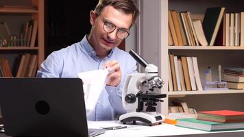 Close-up portrait a frustrated scientist is crumpling a piece of paper while working with a microscope and laptop in his office, surrounded by bookshelves filled with scientific literature video