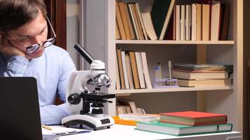 Close-up portrait young male scientist falling asleep at his desk while working late at night in his office or laboratory, he is surrounded by books and scientific instruments video