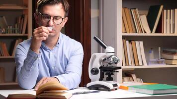 Close-up portrait young male scientist wearing glasses, smelling a chemical sample while taking notes in a laboratory filled with microscopes, books, and test tubes, engaged in research and analysis video