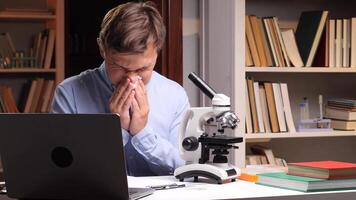Close-up portrait scientist blowing his nose while working at a cluttered laboratory desk, surrounded by a microscope, laptop, and books video