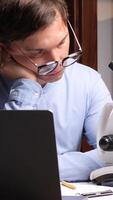 A man with glasses sitting at a desk with a laptop and microscope video