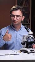 A man in glasses is holding a microscope and giving a thumbs up video