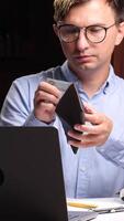 A man with glasses holding a wallet and looking at his laptop video