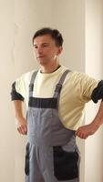 Vertical construction worker wearing overalls standing with hands on his hips in an unfinished room, looking to the side with a serious expression video